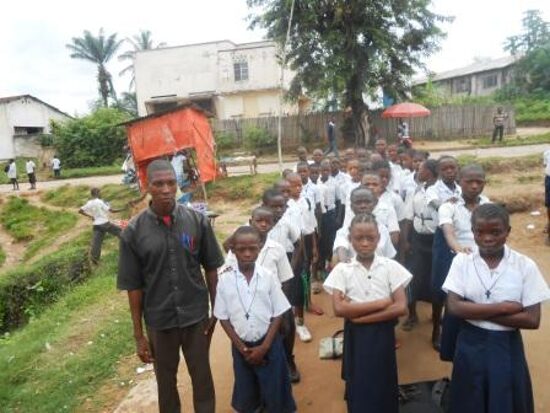 Pupils in white and blue uniforms lined up in rows in the schoolyard with their teachers