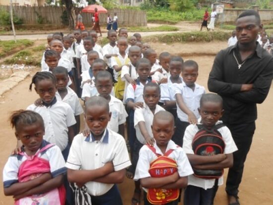 Pupils in white and blue uniforms lined up in rows in the schoolyard with their teachers