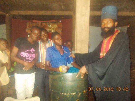 Father Theophile baptizing by immersion in a large barrel font, the faithful looking on