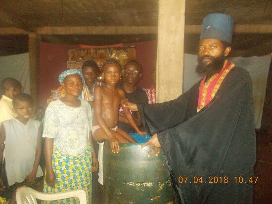 Father Theophile baptizing by immersion in a large barrel font, the faithful looking on
