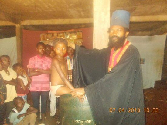 Father Theophile baptizing by immersion in a large barrel font, the faithful looking on