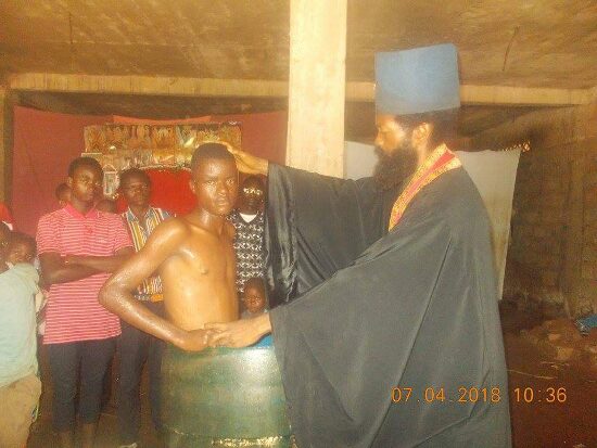 Father Theophile baptizing by immersion in a large barrel font, the faithful looking on