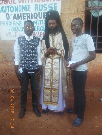 Father Theophile in vestments with newly baptized and their families in front of the church sign of the Parish of the Dormition in Tshikapa