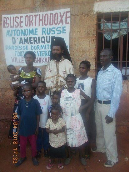 Father Theophile in vestments with newly baptized and their families in front of the church sign of the Parish of the Dormition in Tshikapa