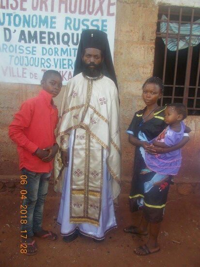 Father Theophile in vestments with newly baptized and their families in front of the church sign of the Parish of the Dormition in Tshikapa